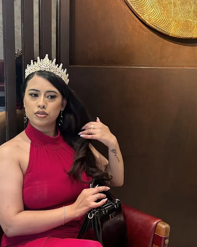 Kay, in a red dress and a crown, seated in a red leather chair against dark wood panelling, one hand in her hair