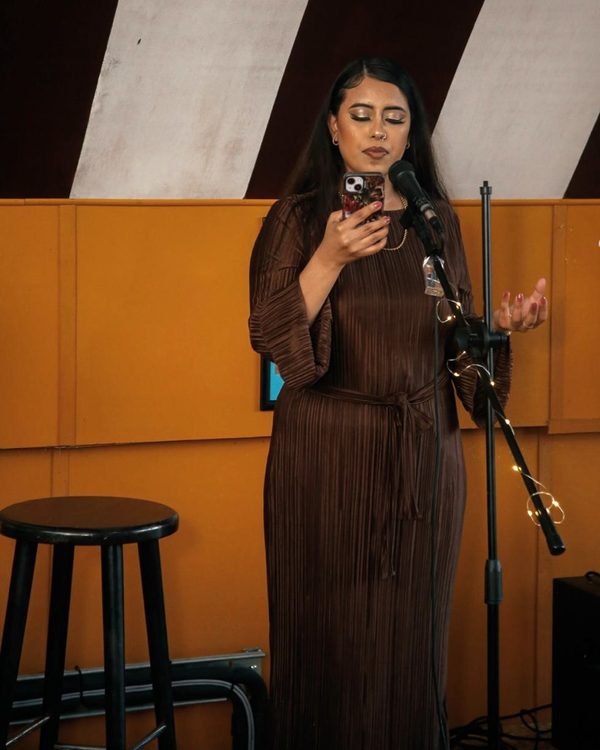 Kay on stage at a microphone, reading from her phone, a stool beside her