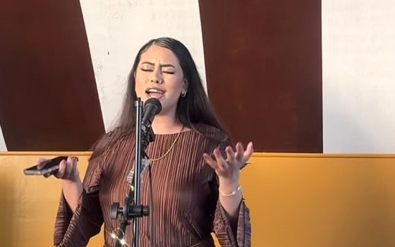 Kay singing on stage at a microphone, both hands open